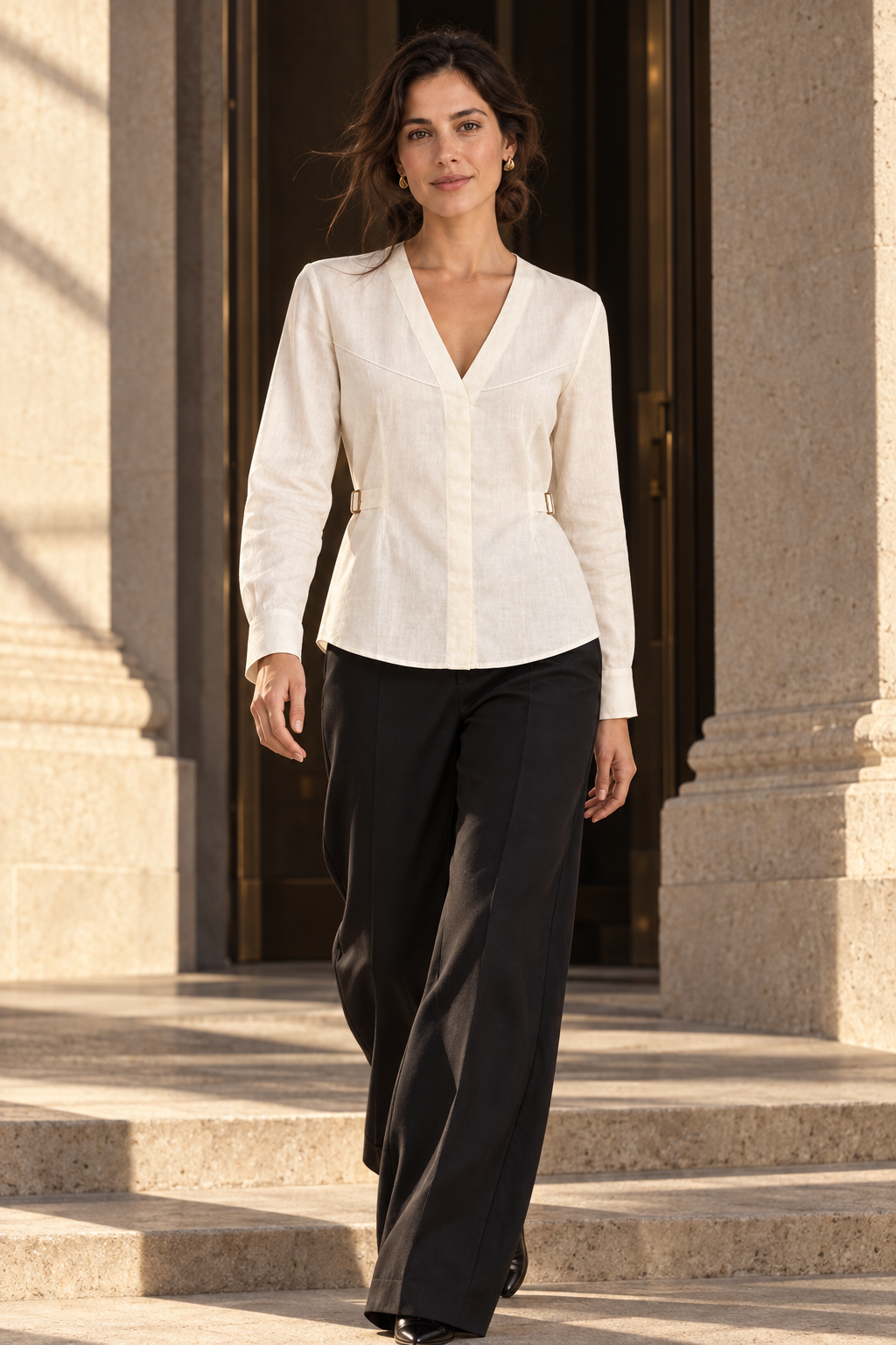 Model stepping through sunlit stone architecture in the ivory V-neck Shirae with curved bust seams, side buckles and black tailored trousers